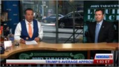 A man sitting at the news desk in front of a sign.